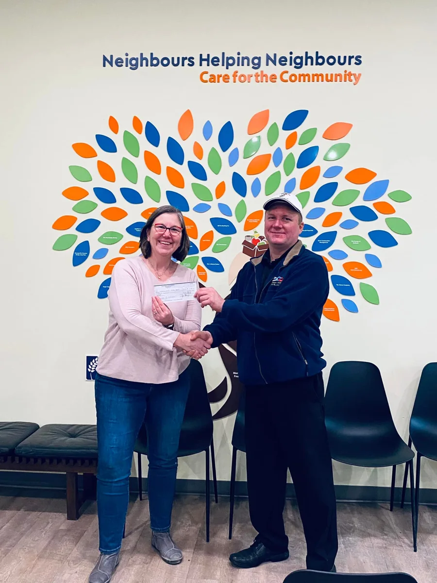 Two community partners shaking hands while presenting a donation cheque beneath a Neighbours Helping Neighbours display