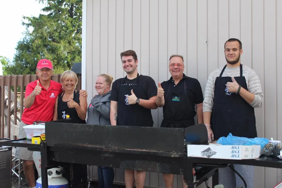 Six volunteers smiling behind an outdoor community cookout grill